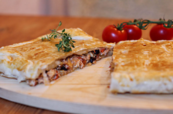 Auf einem Holzbrettchen liegt eine andalusische Blätterteigpastete namens Pastela moruna. Sie ist in der Mitte durchgeschnitten und gibt den Blick frei auf eine Füllung aus Hühnchenfleisch, Mandeln und Rosinen.