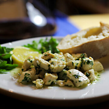 Ein Teller mit gewürfeltem, gebratenen Hühnerfleisch in einer Zitronensauce mit Knoblauch und Petersilie. Im Hintergrund liegt eine Scheibe Ciabatta und eine Deko aus Zitrone und Petersilie.