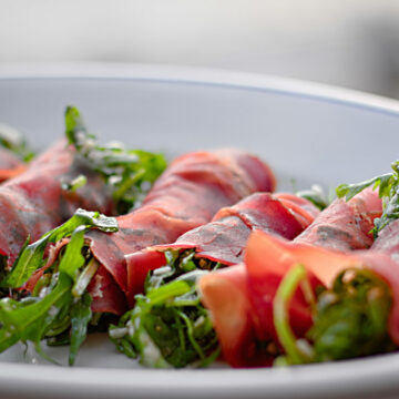 Auf einer Platte liegen sieben mit Rucola gefüllte Bresaola-Röllchen nebeneinander.
