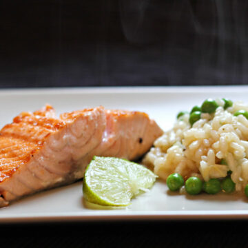 Ein Teller mit noch dampfenden Erbsenrisotto und einem Lachsfilet, garniert mit einer Limettenspalte