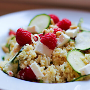 Ein weißer Teller mit einem Couscous-Salat, der mit Zucchinischeiben, Frühlingszwiebeln und vor allem mit Himbeeren und Feta zubereitet ist.