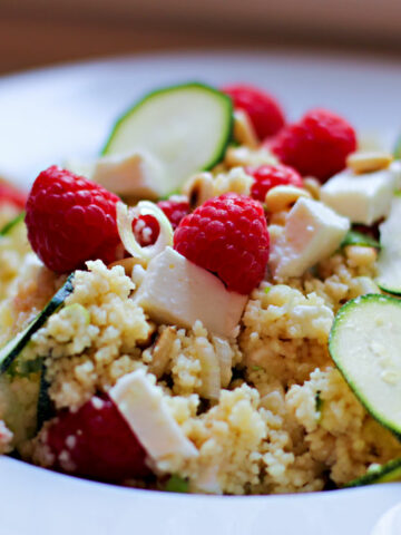 Ein weißer Teller mit einem Couscous-Salat, der mit Zucchinischeiben, Frühlingszwiebeln und vor allem mit Himbeeren und Feta zubereitet ist.
