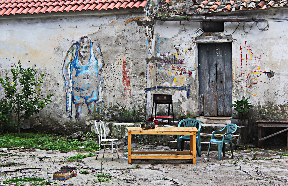 Eine bemalte Hauswand in Agropoli, auf der ein alter Mann in Unterwäsche zu sehen ist. Davoe ein Tisch mit drei Stühlen. Auf dem Tisch sitzt eine Katze.