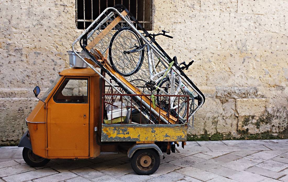 Eine orangene Ape in Lecce, die extrem beladen ist - mit einem Fahrrad, einem Lattenrost und viel Metallschrott.