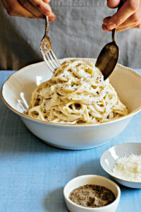 Eine weiße Schüssel mit Spaghetti Cacio e Pepe, die auf einem Tisch mit blauer Tischdecke steht. Jemand richtet sie mit Gabel und Löffel an. Im Vordergrund zwei kleine Schalen mit gemahlenem Pfeffer und Cacio-Käse. (Foto: Kristina Gill)