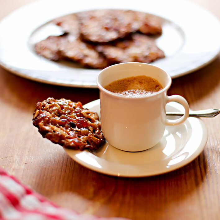 Eine Tasse Espresso mit einem Florentiner Plätzchen, das auf der Untertasse liegt. Im Hintergrund ein Teller mit weiteren Florentinern