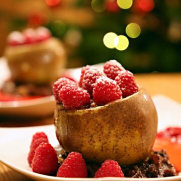 Auf einem Teller steht eine gefüllte Birne in einer Basis aus Schoko-Birnen-Keks-Mischung, verziert mit Himbeeren. Auch die Birne ist an der Schnittfläche mit Himbeeren verziert und mit Puderzucker bestäubt.