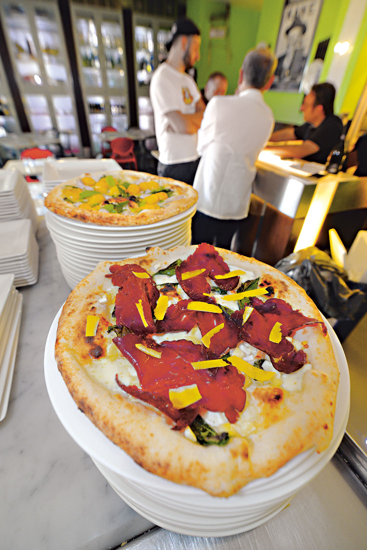 Auf einem Stapel Teller liegt eine Pizza mit Zitrone, im Hintergrund sind Kellner und Restaurantbesucher zu sehen.