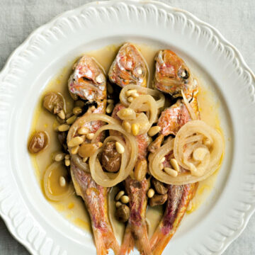 Ein weißer Teller, auf dem drei Rotbarben in Sud angerichtet und mit Zwiebeln und Pinienkernen garniert sind. (Foto: Kristina Gill)