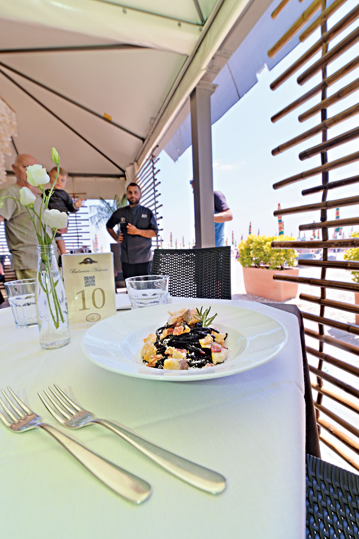 In einem sonnigen Strandrestaurant steht auf einem Tisch ein Teller frische Pasta mit Stockfisch, Pancetta und Schäfskäse
