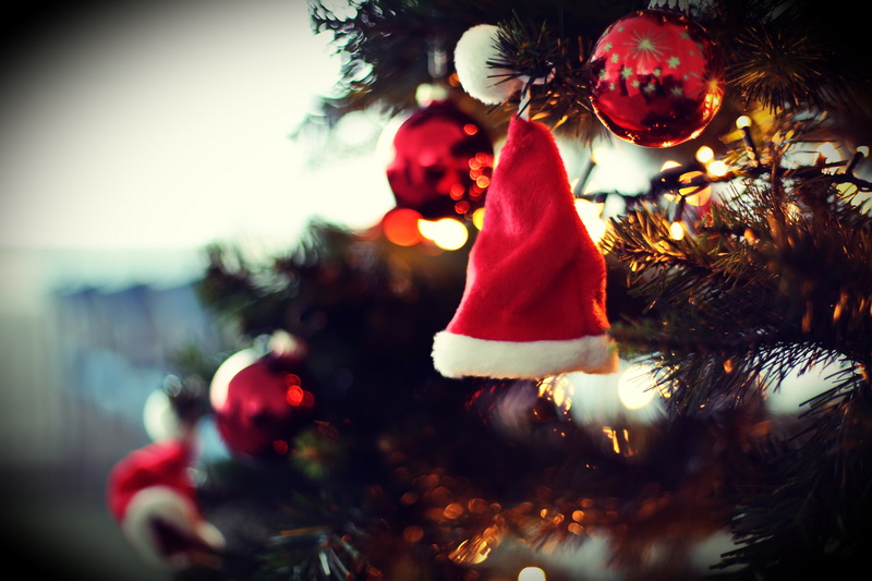 Ein Tannenzweig vom Weihnachtsbaum, an dem im Hintergrund rote Glaskugeln hängen und im Vordergrund eine kleine Weihnachtsmann-Mütze