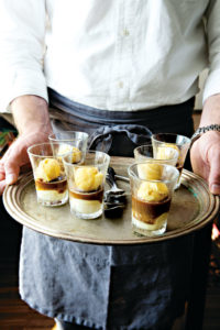 Ein Kellner zeigt ein Tablett, auf dem sechs kleine Serviergläser stehen. Sie sind mit Zabaione-Eis in Kaffeesauce gefüllt. In der Mitte des Tabletts liegen Dessertlöffel. (Foto: Kristina Gill)