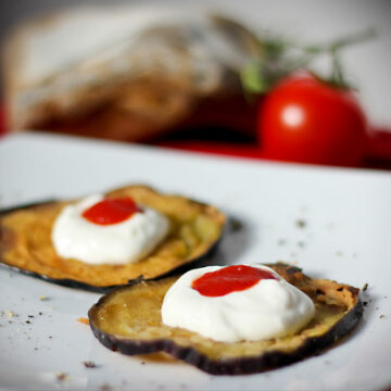 Ein Teller mit zwei frittierten Auberginenscheiben, die jeweils mit einem etwas Knoblauch-Joghurt und einem Klecks Tomatensauce angerichtet sind.