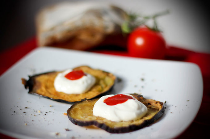 Ein Teller mit zwei frittierten Auberginenscheiben, die jeweils mit einem etwas Knoblauch-Joghurt und einem Klecks Tomatensauce angerichtet sind.