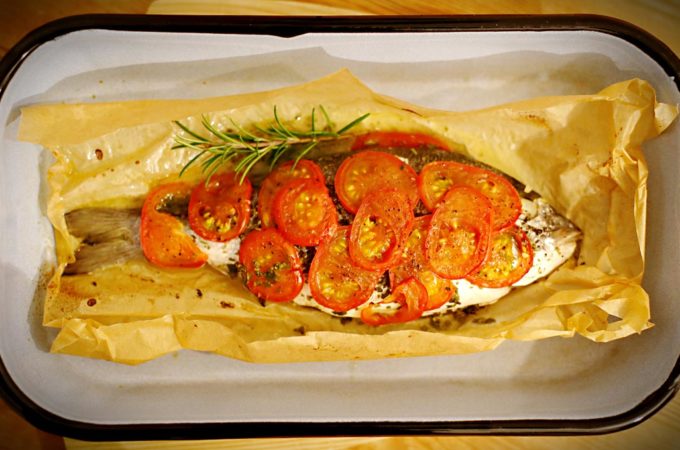 In einer Backofenform liegt eine Dorade, die mit Tomatenscheiben und Kräutern belegt ist.