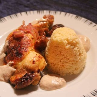 Auf einem Teller liegt eine gegarte Hähnchenkeule mit Mandelsauce und Feigen und aufgetürmtem Couscous.