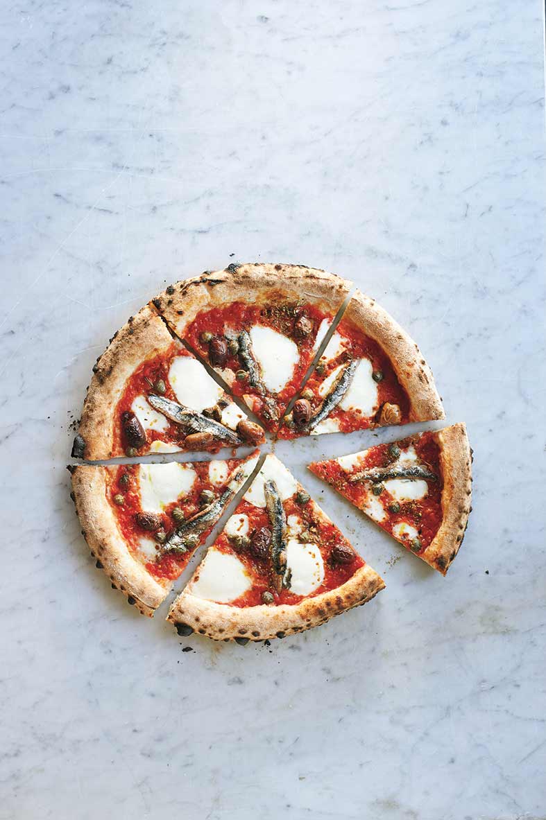 Eine runde Pizza, die in acht Stücke geschnitten wurde. Sie ist belegt mit Sardellenfilets, Tomatensauce, Mozzarella, Oliven und Kapern.