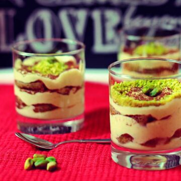 Auf einer roten Decke stehen drei Gläser mit Pistazientiramisu, die mit gehackten Pistazien dekoriert sind.