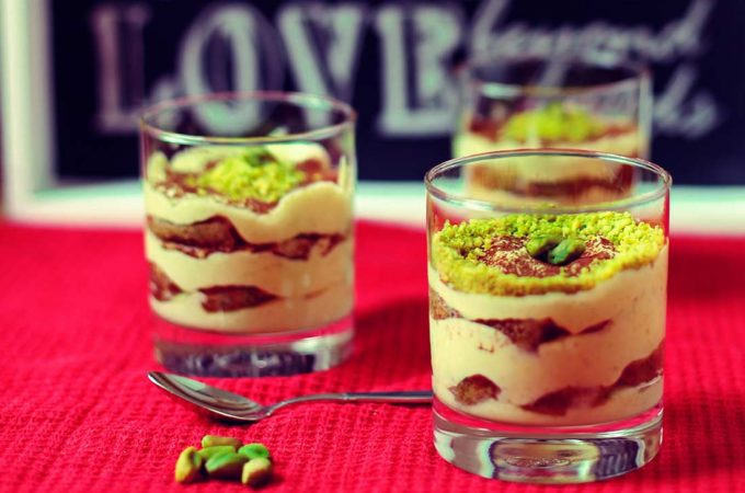 Auf einer roten Decke stehen drei Gläser mit Pistazientiramisu, die mit gehackten Pistazien dekoriert sind.