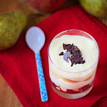 Ein Dessertglas mit Birnen-Tiramisu auf einer roten Decke, daneben eine Birne und ein Löffel.