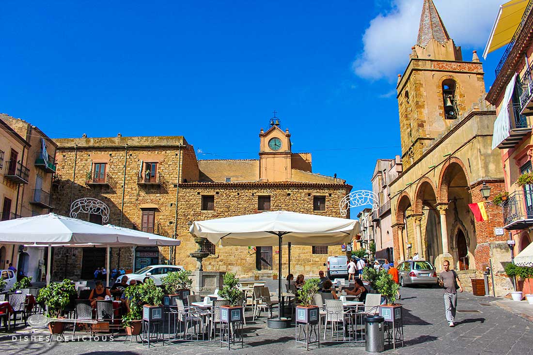 Piazza Margherita in Castelbuono: mitten auf dem Platz stehen Tische eines Cafès, rundherum alte Bauten wie die Chiesa Matrice Vecchia.