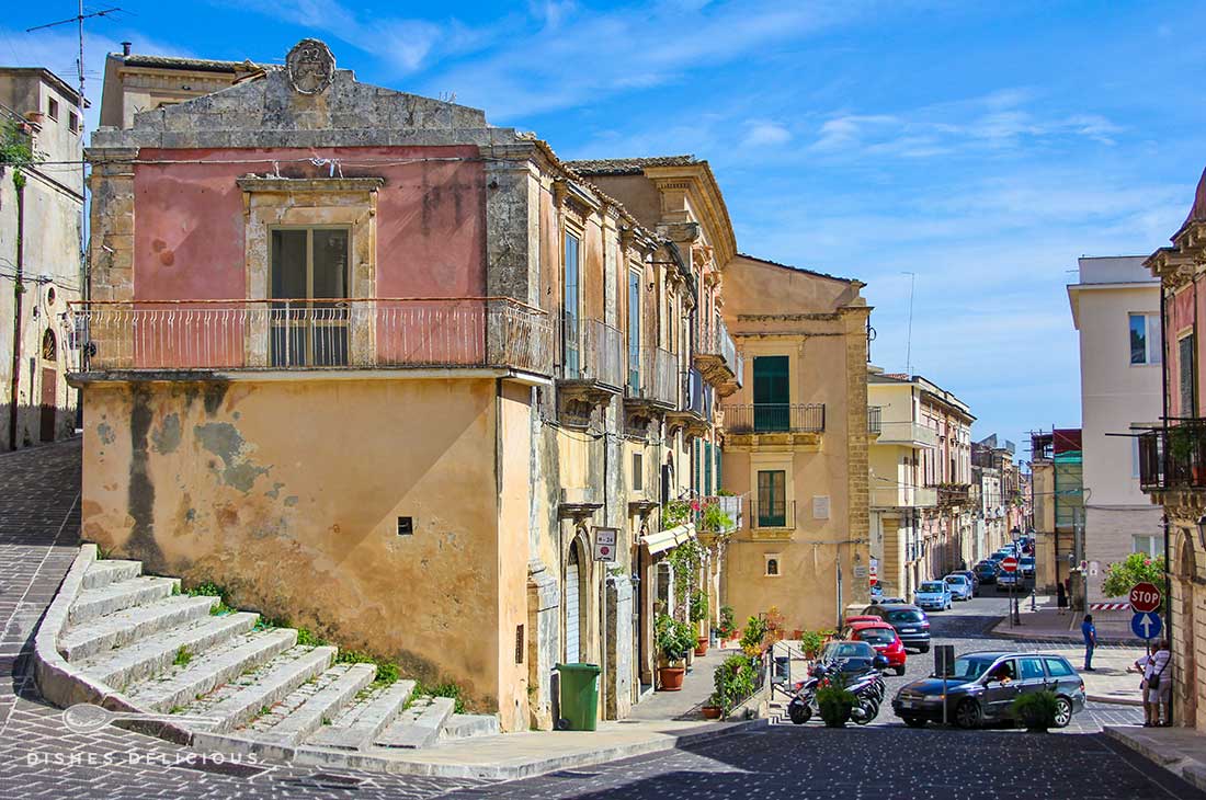 Eine Straße in Palazzolo Acreide, im Vordergrund ein altes Haus, an dem eine Treppe entlangführt.