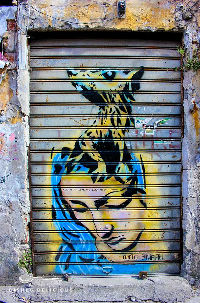 Street-Art: An die Jalousie eines Ladens ist eine Maria gesprüht, aus deren Kopf eine Ratte wächst.