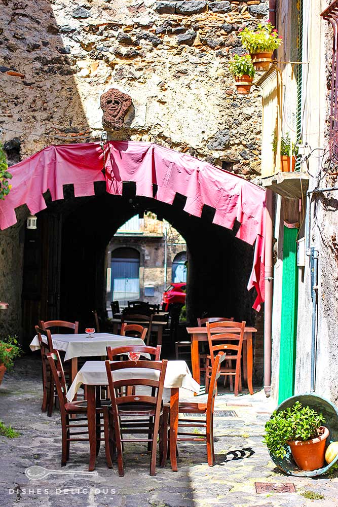 Osteria-Tische und Stühle vor einer dekorierten Unterführung in der Altstadt von Randazzo.