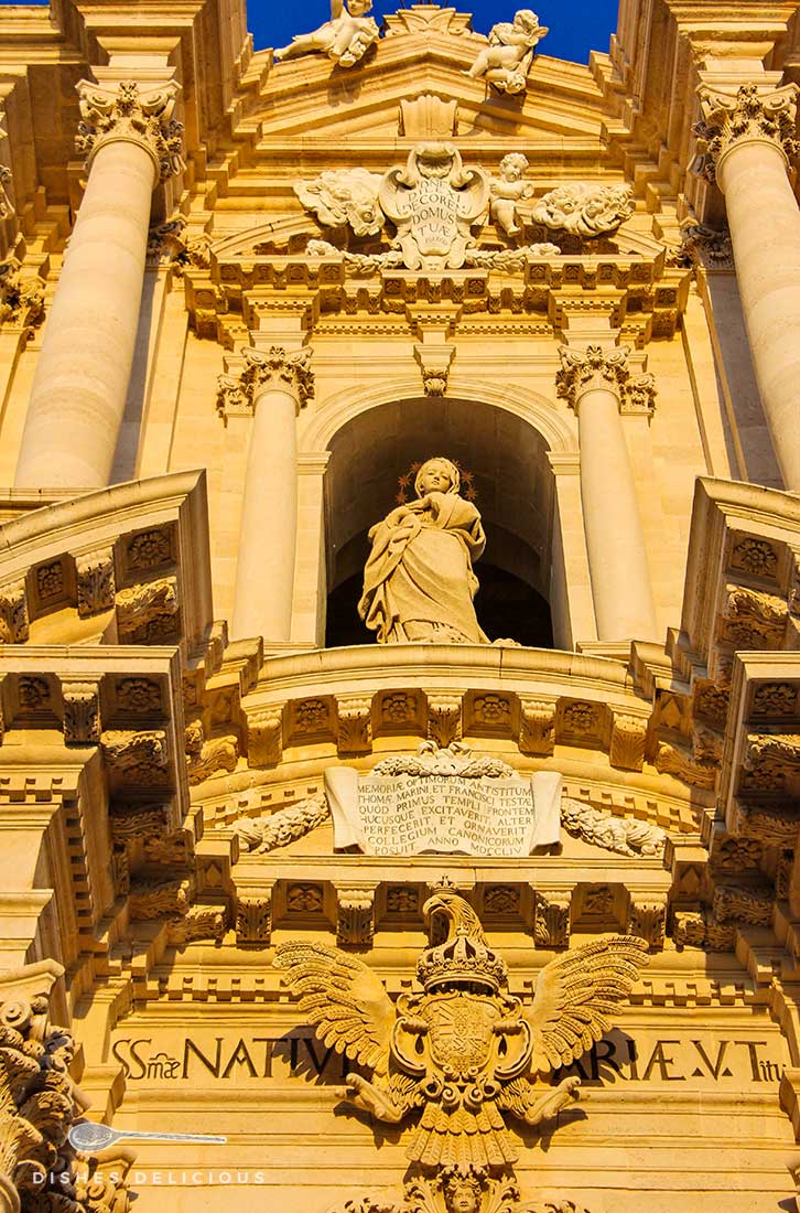 Ausschnitt aus der Barockfassade des Doms von Syrakus, in der Mitte ist zwischen Säulen eine Maria zu sehen, am Rande viele Engelsverzierungen.