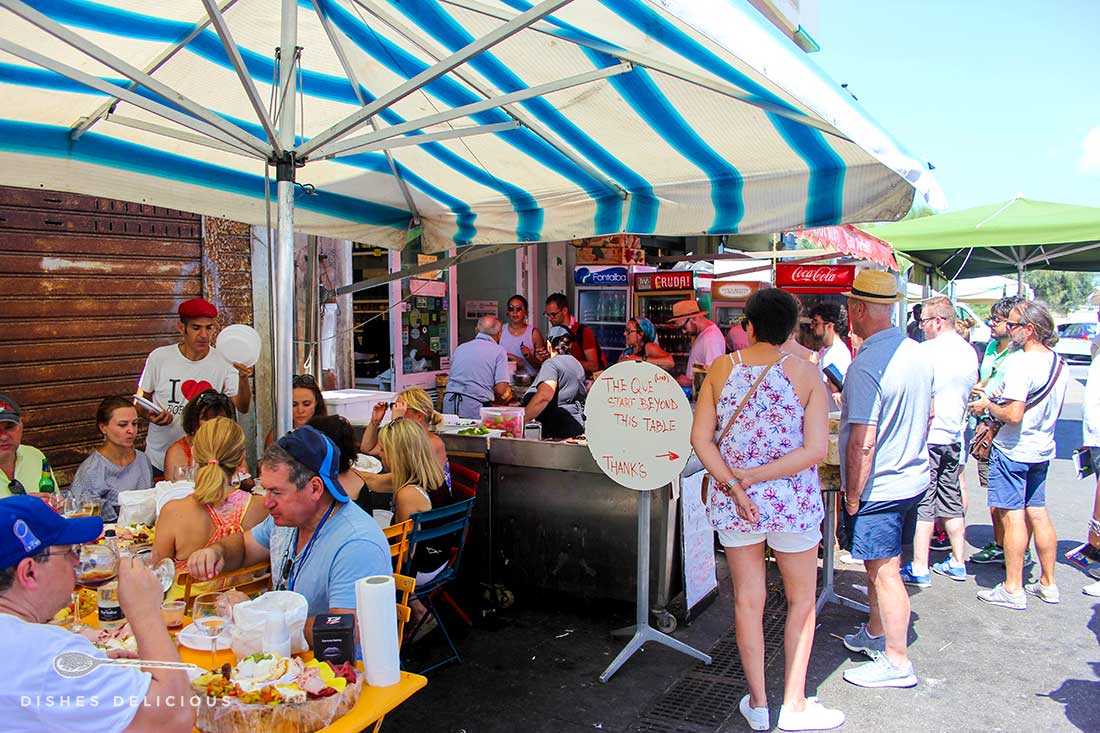 Warteschlange vor dem Caseficio Borderi. Auf der linken Seite vor der Käserei stehen Tische, an denen die Gäste Antipasti-Platten essen.