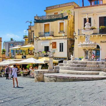 Die Piazza Duomo in Taormina. Vor einigen Cafès mit Tischen im Freien steht ein großer Brunnen.