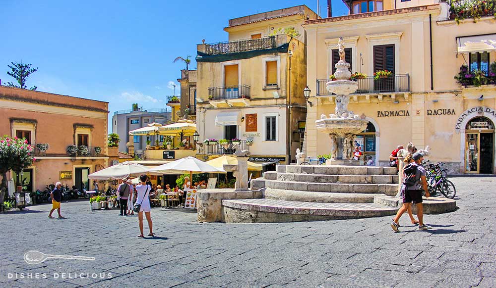Die Piazza Duomo in Taormina. Vor einigen Cafès mit Tischen im Freien steht ein großer Brunnen.