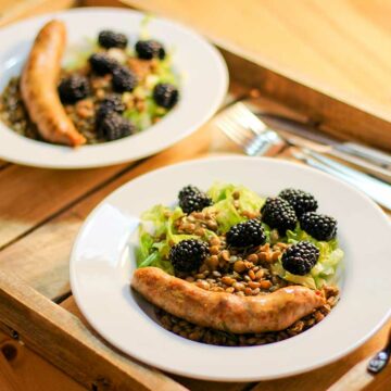Zwei Teller, auf denen Linsen, Brombeeren, Salat und eine Salsiccia liegen. Die Teller stehen auf einem Holzbrett.