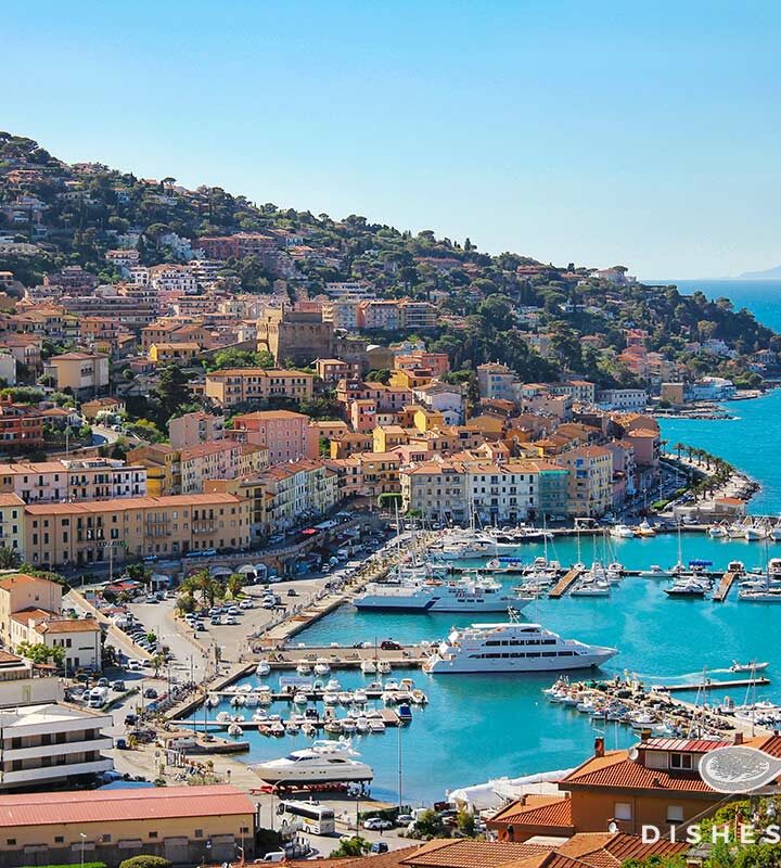Panoramafoto von Porto Santo Stefano, das sich am Hang entlangzieht, im Vordergrund der Hafen.