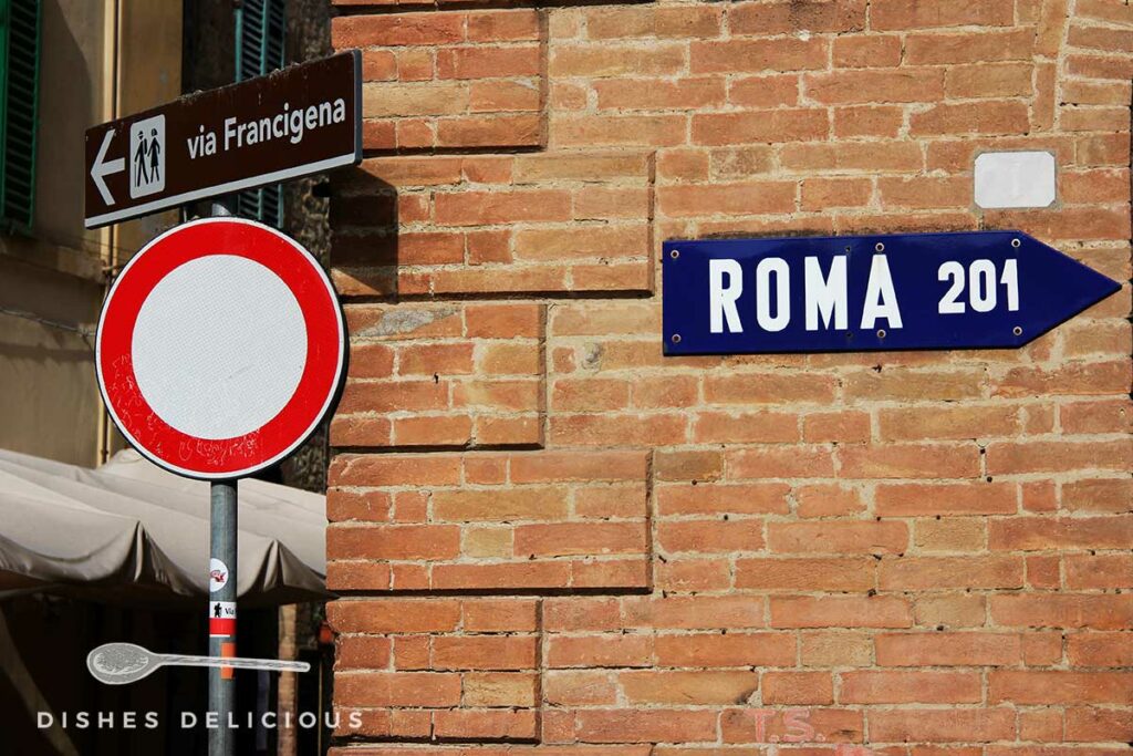 Foto von mehreren Straßenschildern an einem Backsteinbau. Ein Schild weist in Richtung Rom