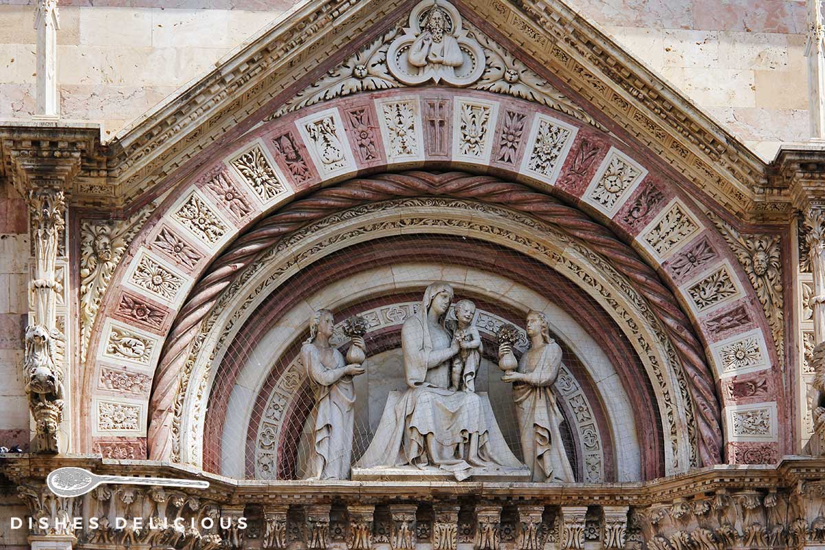 Foto einer Steinverzierung am Dom von Grosseto, die Maria mit Jesus zwischen zwei Heiligen zeigt.