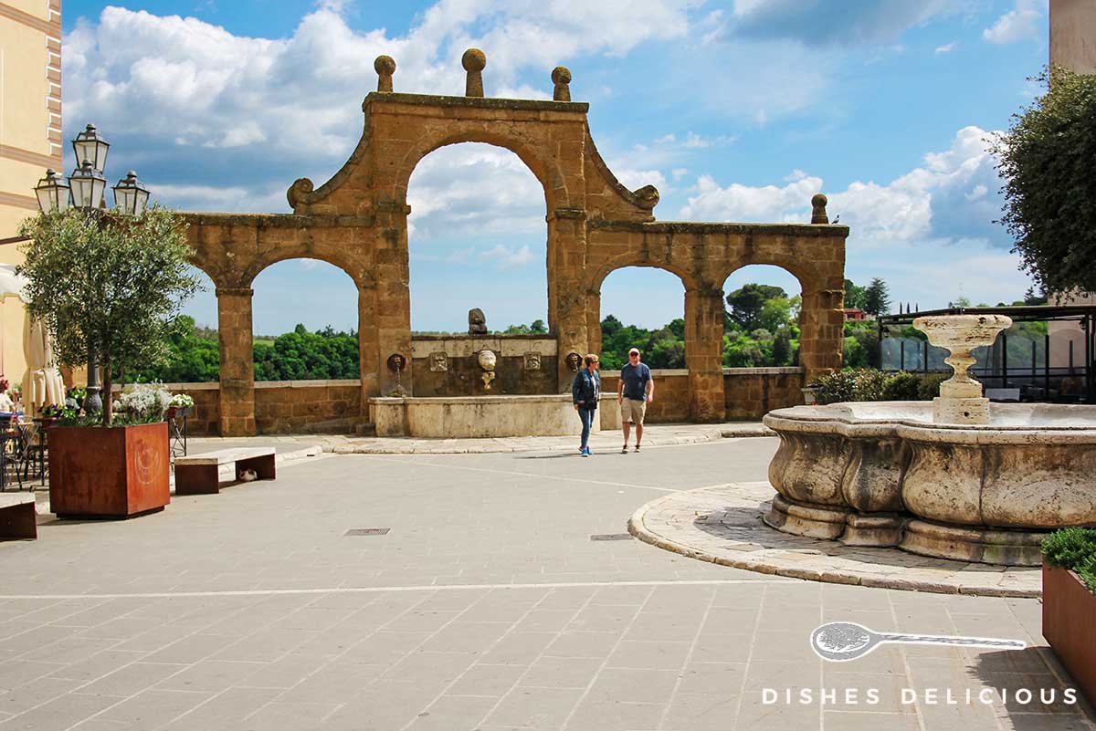 Foto eines Platzes, im Vordergrund ein alter Rundbrunnen, im Hintergrund ein weiterer Brunnen mit fünf Bögen..