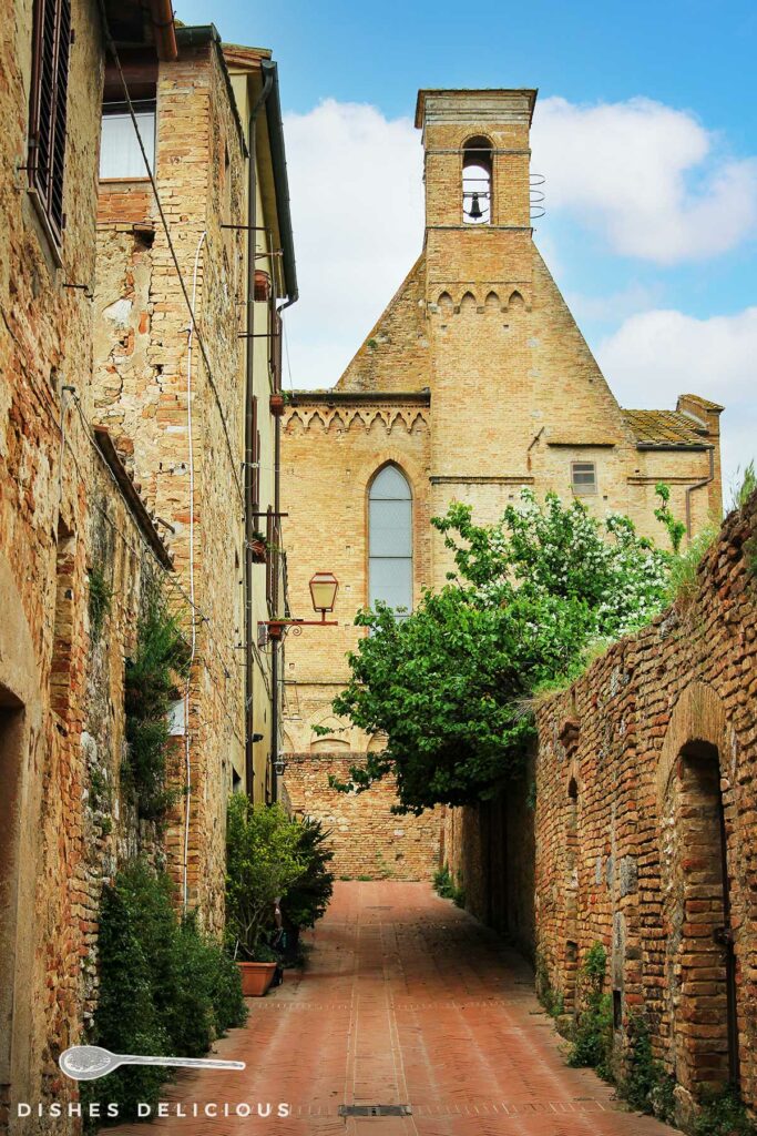 Foto von einer Gasse an der Stadtmauer von San Gimignano, im Hintergrund steht eine Kirche.