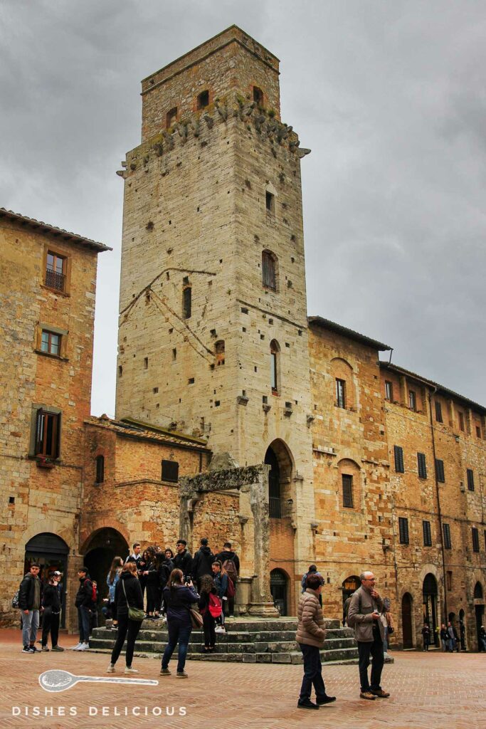 Foto vom hohen Turm Torre del Diavolo, vor dem ein alter Brunnen und viele Touristen stehen.