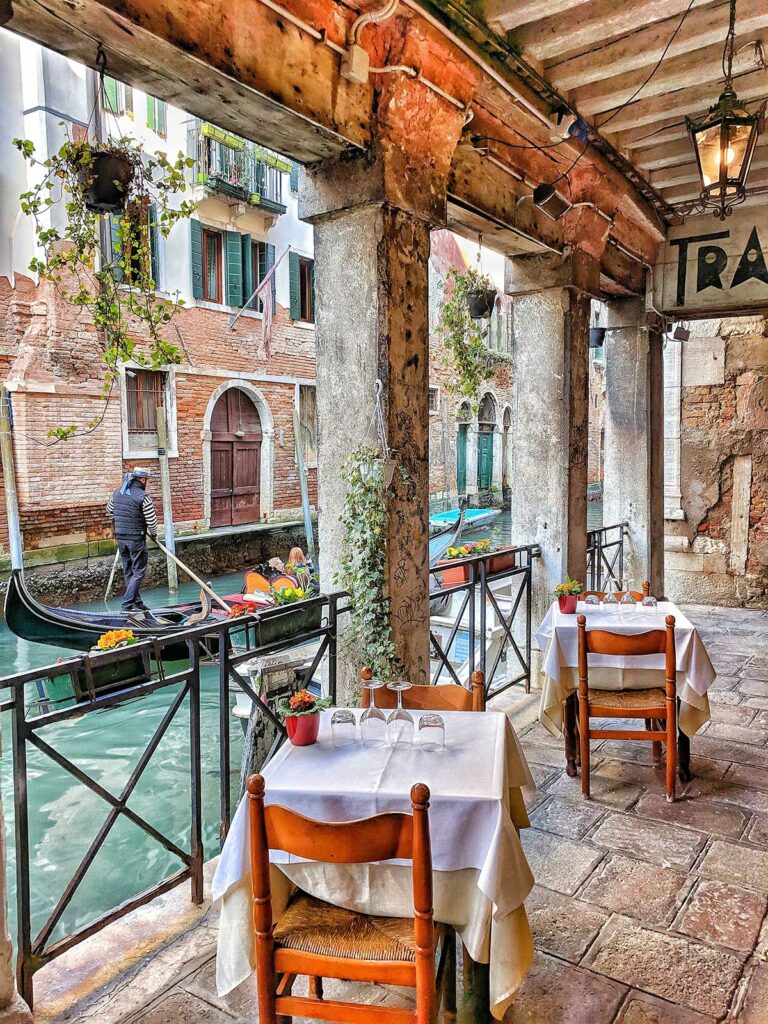 Gedeckte Osteria-Tische an einem Kanal in Venedig, eine Gondel fährt vorbei.