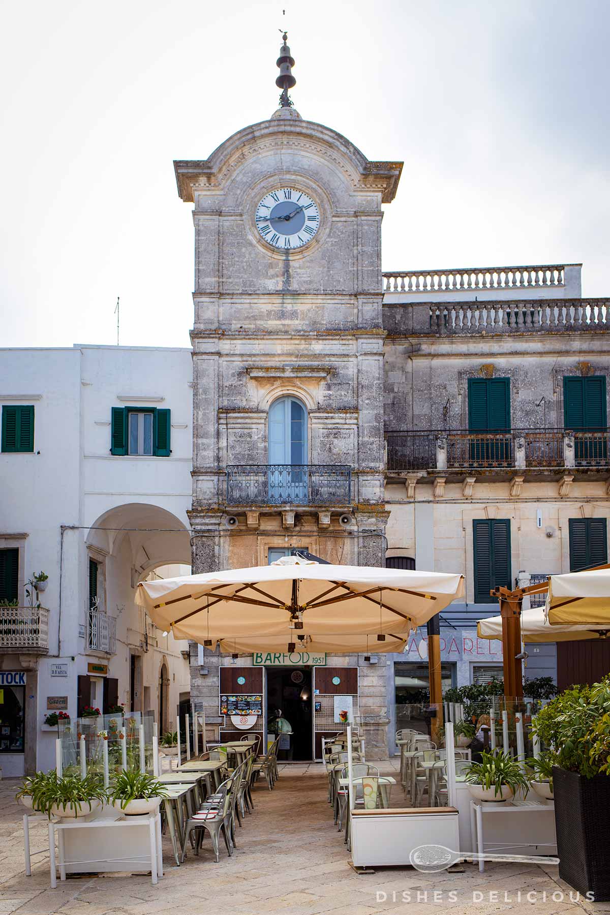 Alter Uhrenturm von Cisternino, vor dem ein Cafè Tische aufgebaut hat.