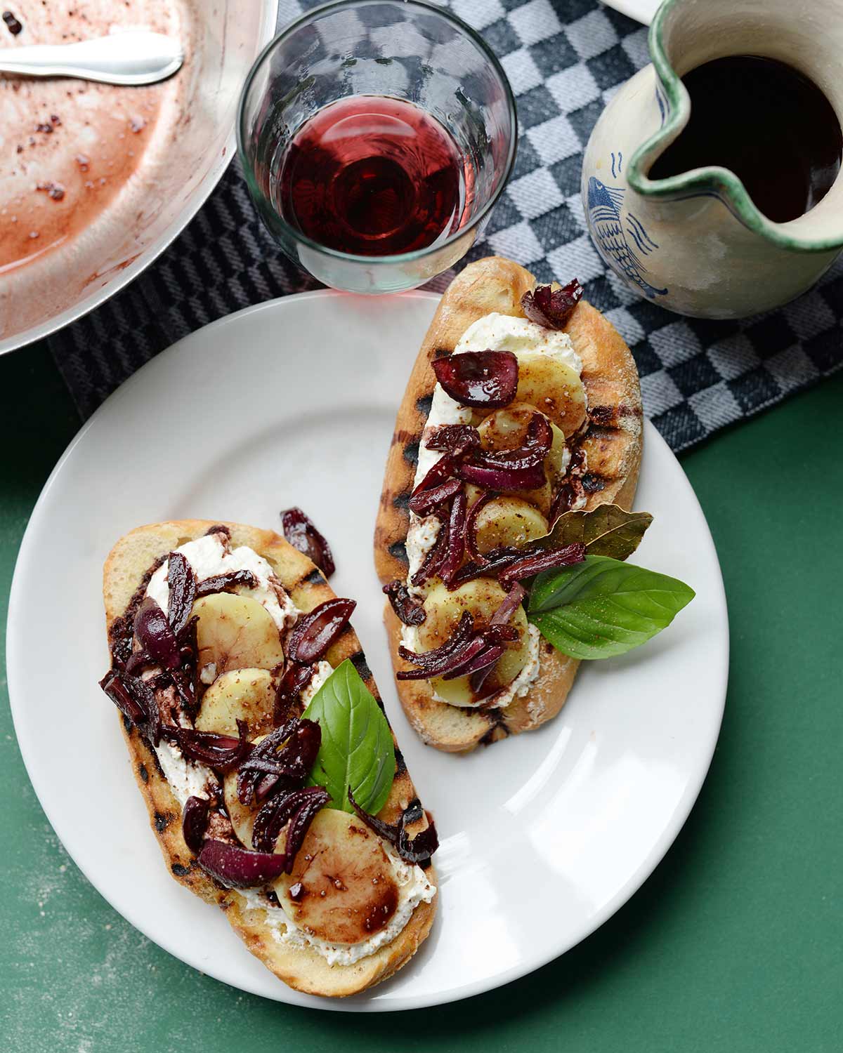 Zwei Crostini mit Ziegenkäse, Kartoffeln und Zwiebeln auf einem Teller, daneben ein Krug und Glas mit Wein.