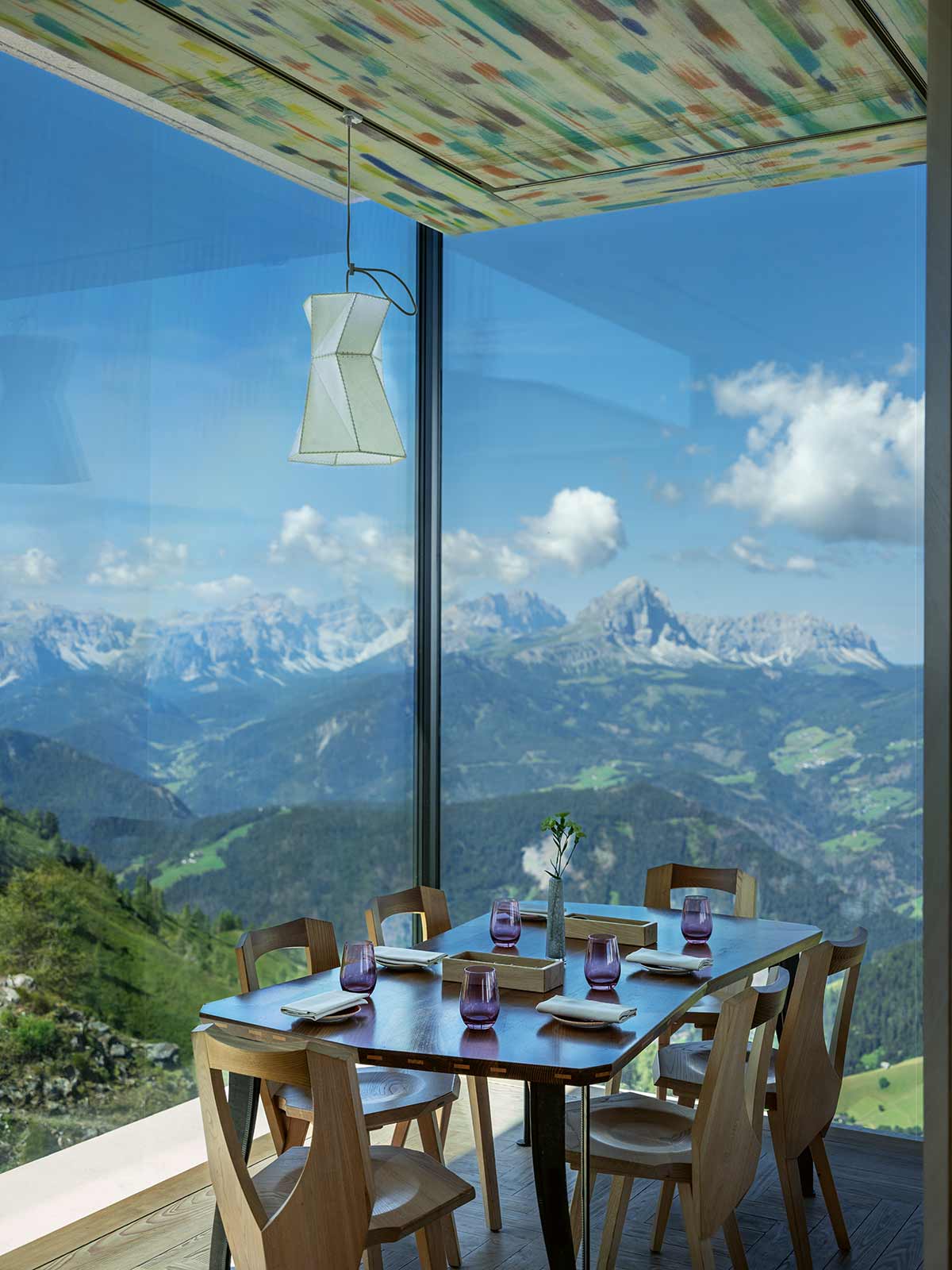 Ein gedeckter, moderner Holztisch in einem Restaurant. Dahinter breit verglaste Fenster mit spektakulärem Blick auf die Alpenlandschaft.