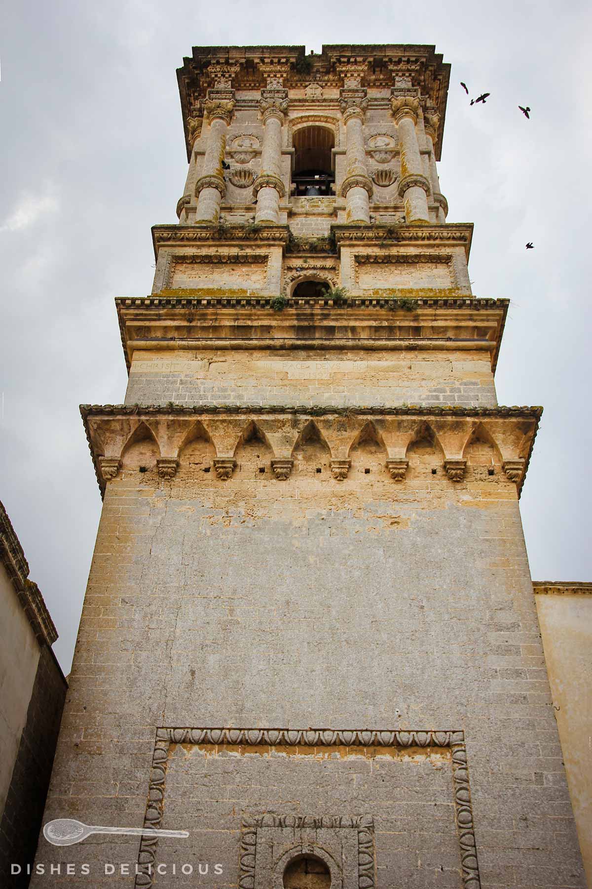 Der Torre Campanaria der Basilica von Copertino, der im oberen Abschnitt mit Säulen-Elementen verziert ist. Vögel kreisen um den Turm.