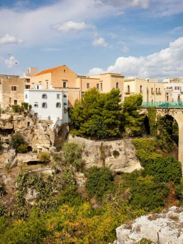Teilansicht der Altstadt von Massafra, die auf einem Felsen liegt. Eine Brücke über einer mit Büschen und Bäumen bewachsenen Schlucht führt in die Altstadt.