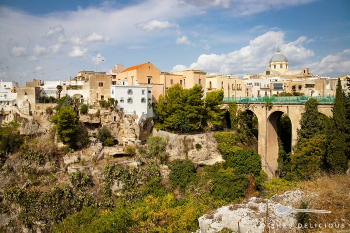 Teilansicht der Altstadt von Massafra, die auf einem Felsen liegt. Eine Brücke über einer mit Büschen und Bäumen bewachsenen Schlucht führt in die Altstadt.