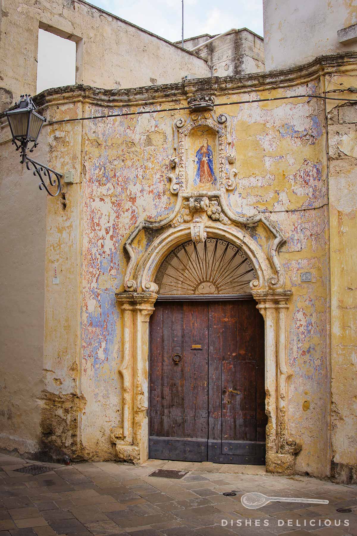 Barockverziertes Eingangstor zu einem Palazzo, das Mauerwerk bröckelt, die Farbe der darauf gemalten Fresken blättern ab.