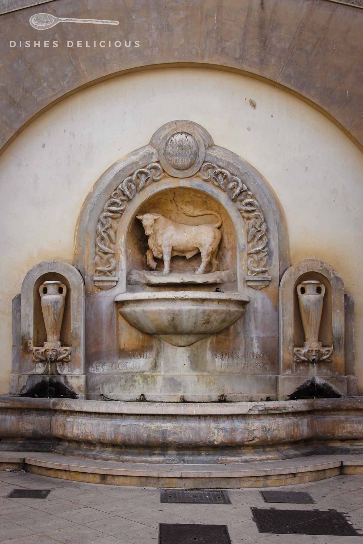 Ein steinener Brunnen mit einer Stierfigur oberhalb der Wasserschale. Rechts und links ist eine Amphore in den Brunnen eingearbeitet.