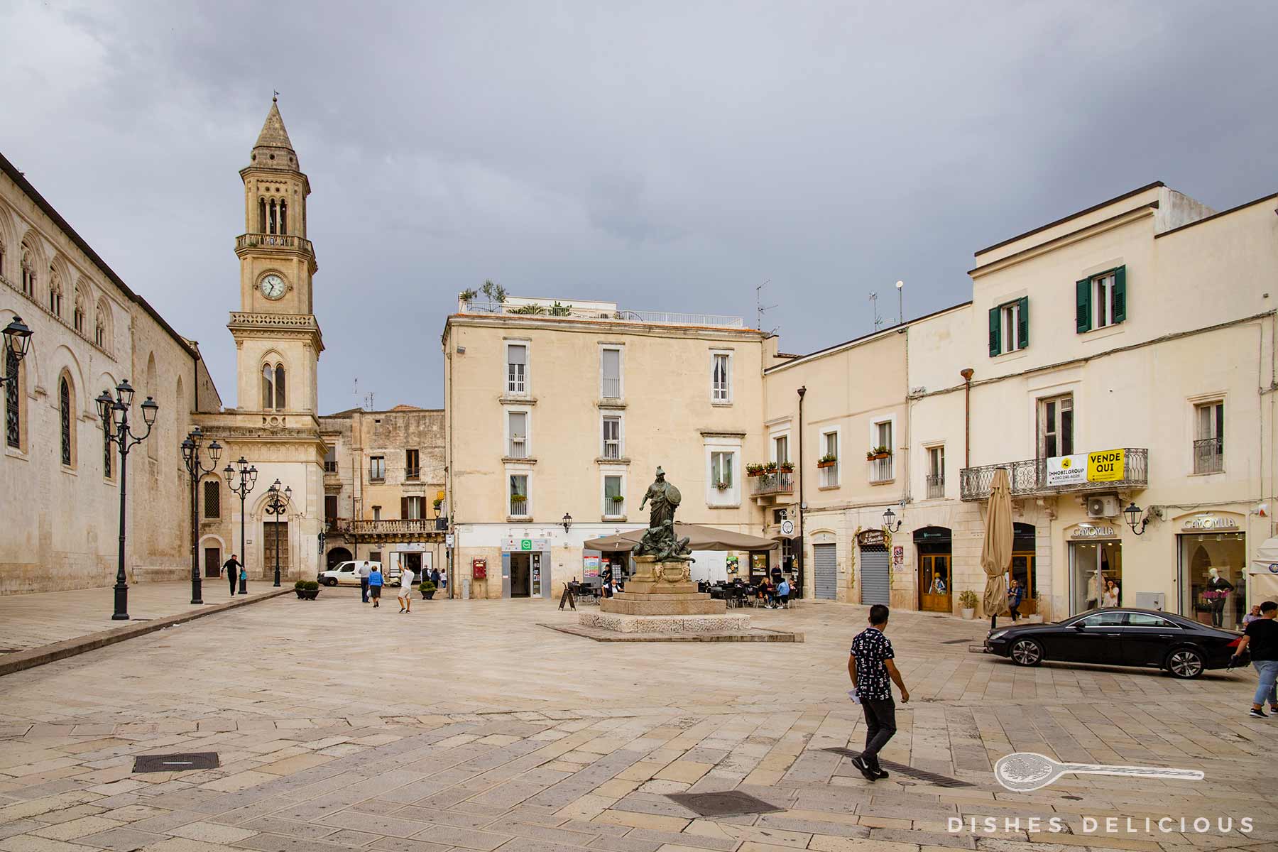 Zentrale Piazza in Altamura, links der Dom und Glockenturm, in der Mitte ein Denkmal, dahinter Geschäftsgebäude und eine Bar.