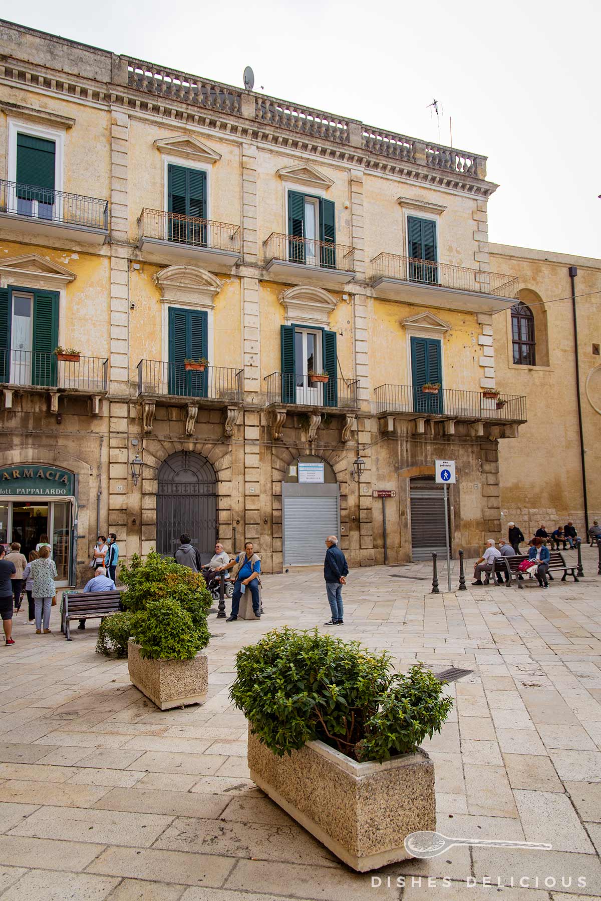 Stattlicher Palazzo an der Piazza Repubblica, davor Pflanzenkübel und Bänke, auf denen Senioren sitzen.